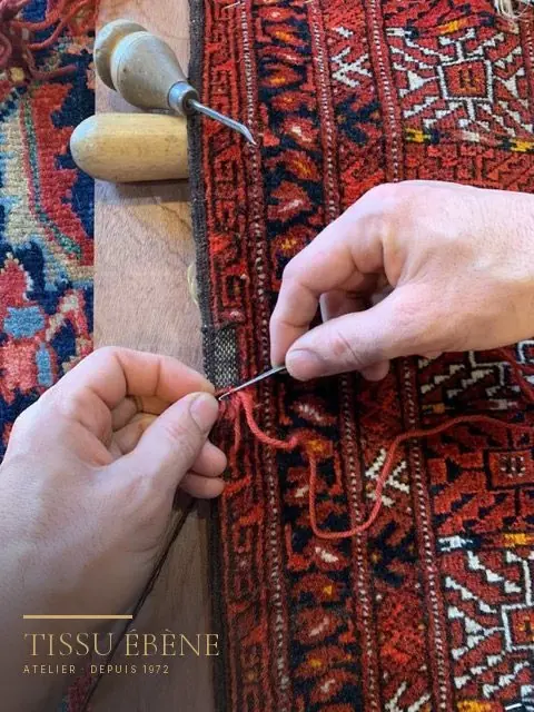 Mains cousant au fil rouge la lisière d&rsquo;un tapis en laine à motifs rouges, outils en bois à côté