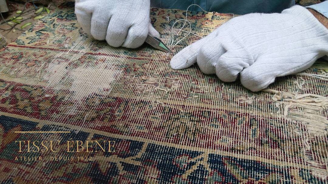 Mains gantées tenant une pince sur un tapis usé jusqu&rsquo;à la trame, décor floral rouge et bleu