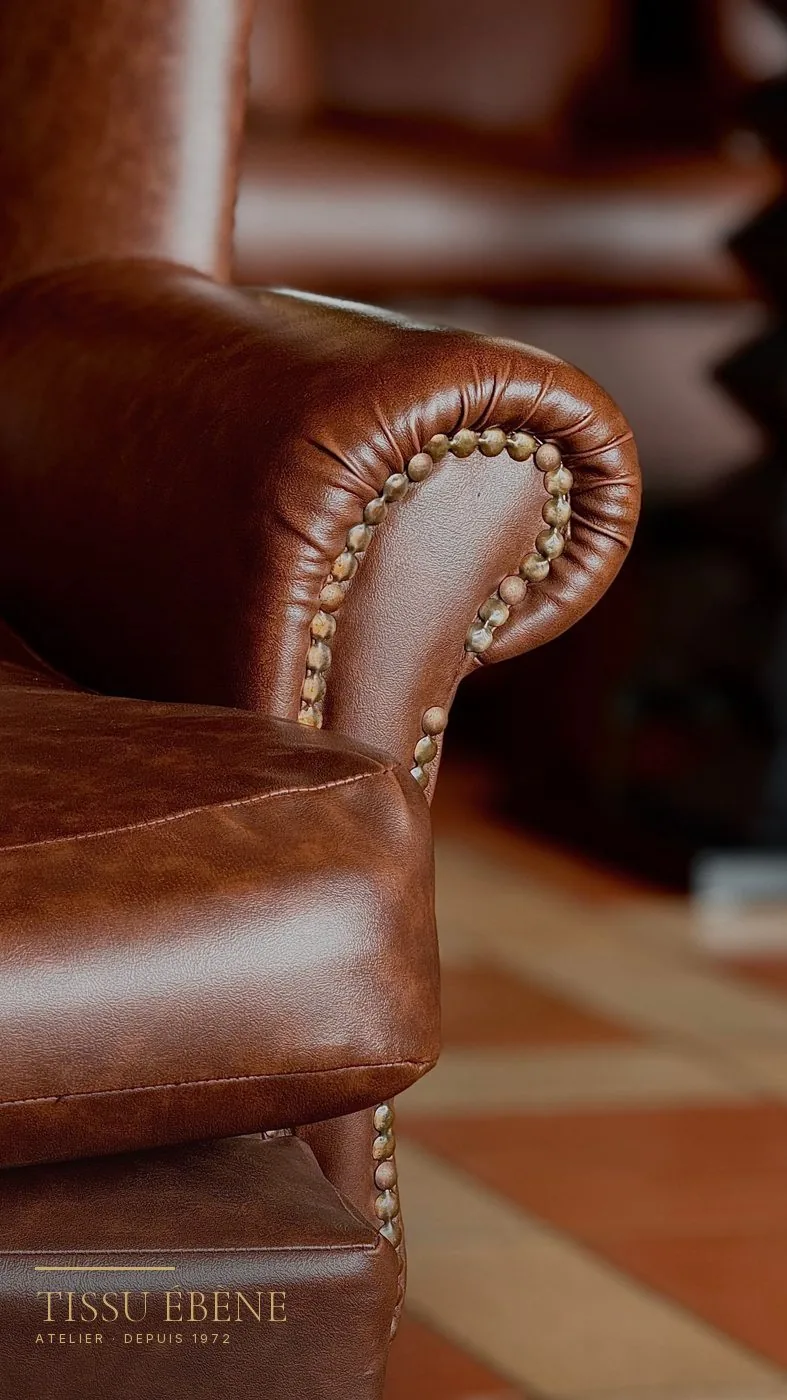 Gros plan sur l’accoudoir d’un fauteuil en cuir brun patiné, clous de tapissier apparents
