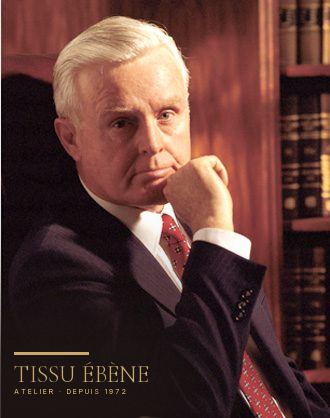 Portrait d&rsquo;un homme aux cheveux blancs en costume sombre et cravate rouge, devant une bibliothèque