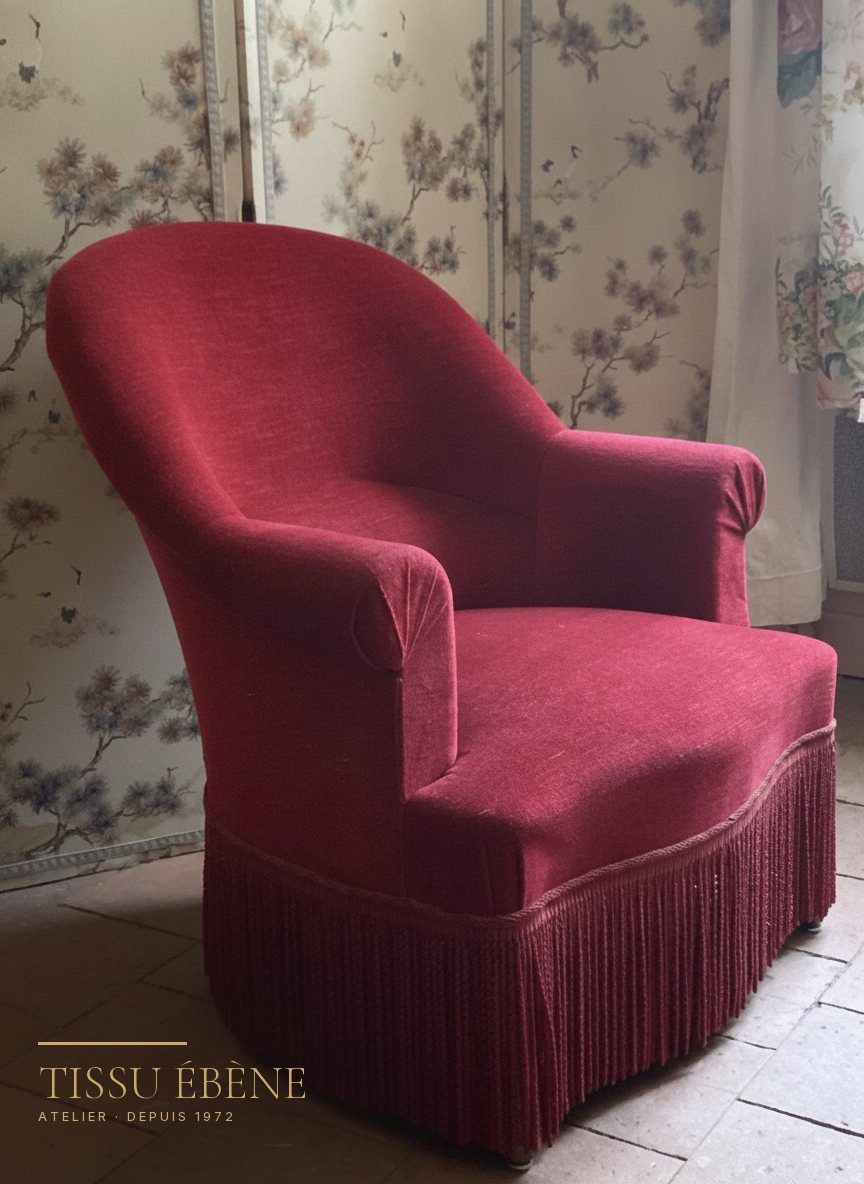 Fauteuil crapaud en velours rouge &agrave; franges devant un papier peint fleuri
