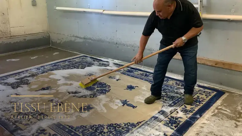 Artisan brossant au balai-brosse un tapis chinois bleu et beige couvert de mousse, au sol